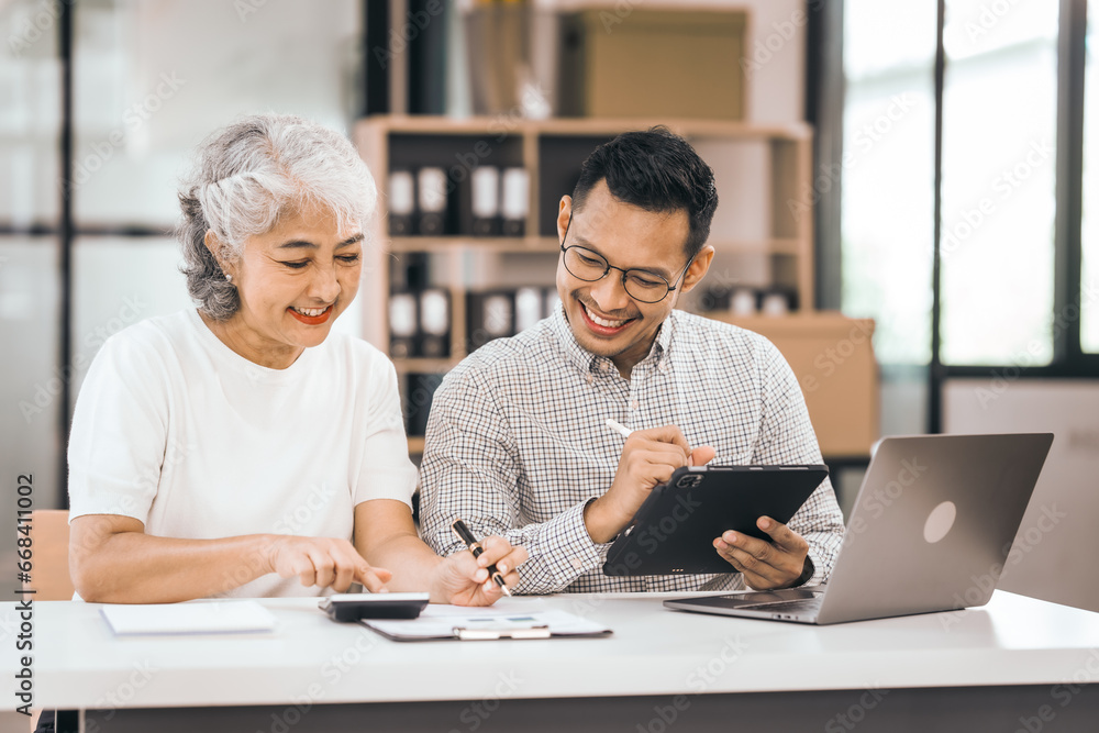 An older mature woman shows something to asian man on a laptop. They smile and work together in an office. They look happy and focused. An older mature woman shows something to asian man on a laptop. They smile and work together in an office. They look happy and focused.