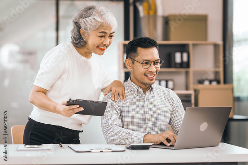 An older mature woman shows something to asian man on a laptop. They smile and work together in an office. They look happy and focused. photo