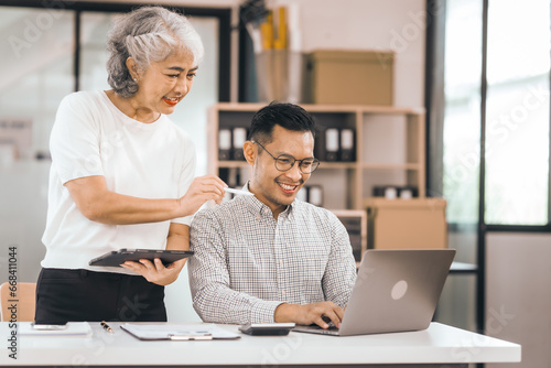 An older mature woman shows something to asian man on a laptop. They smile and work together in an office. They look happy and focused. © makibestphoto