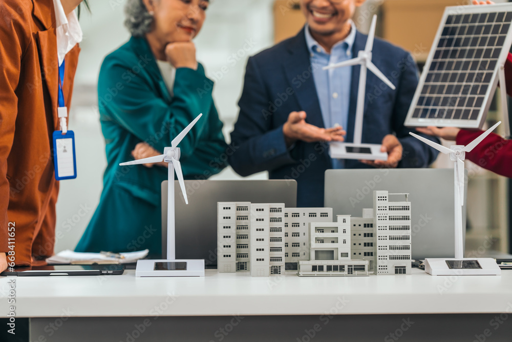 Four asian businesspeople professionals discuss renewable energy in office, showing solar panel and wind turbines with tower building model, apartment. modern business with environmental, clean energy Four asian businesspeople professionals discuss renewable energy in office, showing solar panel and wind turbines with tower building model, apartment. modern business with environmental, clean energy