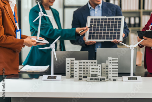 Four asian businesspeople professionals discuss renewable energy in office, showing solar panel and wind turbines with tower building model, apartment. modern business with environmental, clean energy