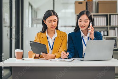 Two young asian business women work in an office. One uses a tablet and looks serious. The other smiles and types on a laptop.