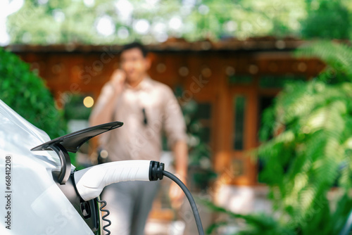 Focused EV electric car recharging at outdoor coffee cafe in springtime garden with blur background of eco friendly man, green city sustainability and environmental friendly EV vehicle. Expedient photo