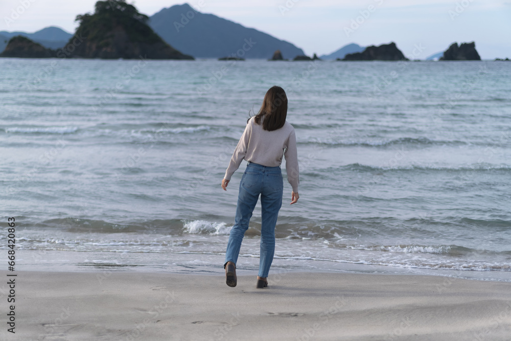  秋の海の砂浜を歩くフレアジーンズをはいた女性（福井県 若狭和田ビーチ）