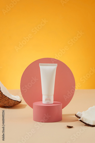 A white cosmetic tube without a label is placed on a pink platform, fresh coconut is decorated around it, the background is divided into two colors. Minimal styling scene with coconut, still life.