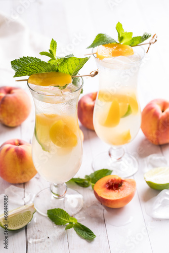 Two peach mojito cocktails surrounded by peaches against a light background.