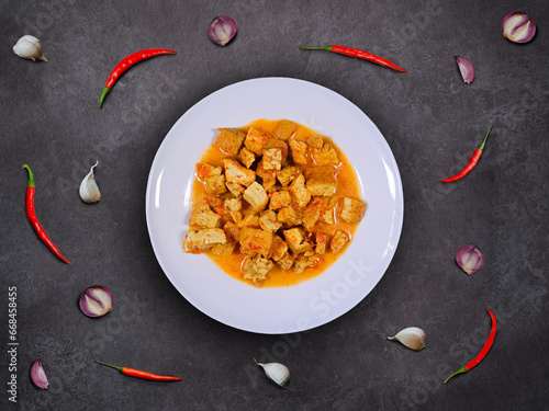 Spicy tempeh and tofu stew in coconut milk with garlics, shallots, and red chilis scattered around the plate. Tempe and tofu curry on a white plate with dark background. Indonesian homemade cuisine. photo