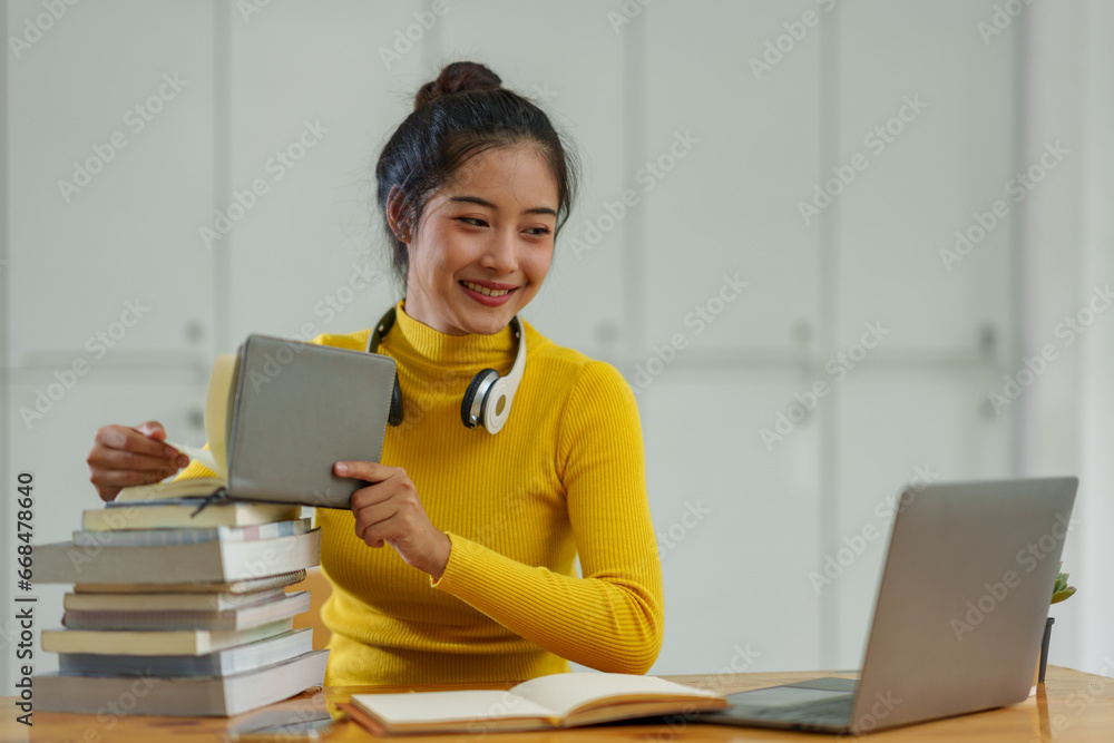Confident Asian female student wearing wireless headphones studying online, searching for information, thinking analytically from documents, textbooks, laptop computer for business studies. Confident Asian female student wearing wireless headphones studying online, searching for information, thinking analytically from documents, textbooks, laptop computer for business studies.