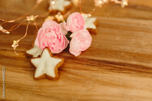 Gingerbread cookies in the shape of stars, merengue cookies and pink roses on a wooden table against a background of bokeh of New Year’s lights. Christmas background. Winter card. Delicious pastries