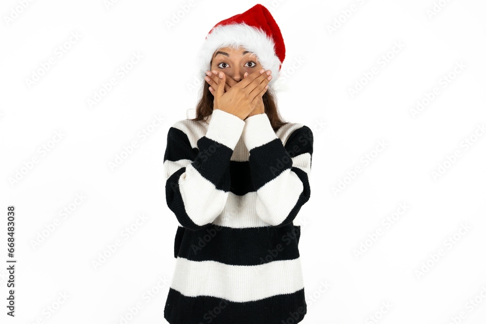 Beautiful girl wearing striped sweater and Santa hat shocked covering mouth with hands for mistake. Secret concept. Beautiful girl wearing striped sweater and Santa hat shocked covering mouth with hands for mistake. Secret concept.