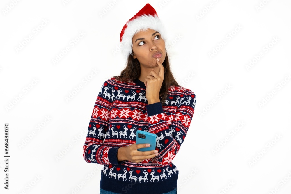 Beautiful girl wearing christmas sweater and Santa hat thinks deeply about something, uses modern mobile phone, tries to made up good message, keeps index finger near lips. Beautiful girl wearing christmas sweater and Santa hat thinks deeply about something, uses modern mobile phone, tries to made up good message, keeps index finger near lips.