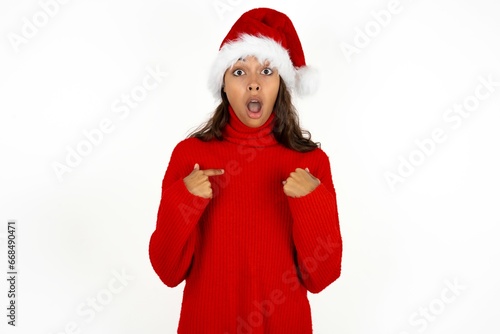 Embarrassed Beautiful woman wearing christmas hat indicates at herself with puzzled expression, being shocked to be chosen to participate in competition, hesitates about something