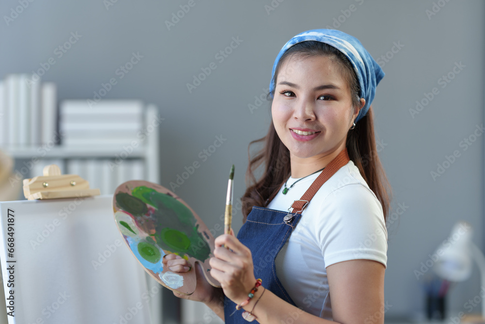Young Asian female artist, painter holds paintbrush and palette with canvas for painting on easel in art studio with acrylic paints on canvas. Lifestyle hobby ideas. Young Asian female artist, painter holds paintbrush and palette with canvas for painting on easel in art studio with acrylic paints on canvas. Lifestyle hobby ideas.