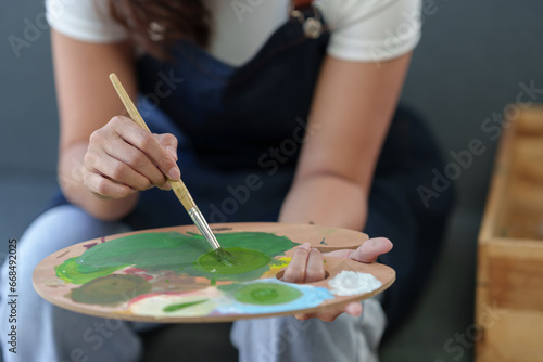 Young Asian female artist, painter holds paintbrush and palette with canvas for painting on easel in art studio with acrylic paints on canvas. Ideas for work ideas hobbies creative lifestyles in art.