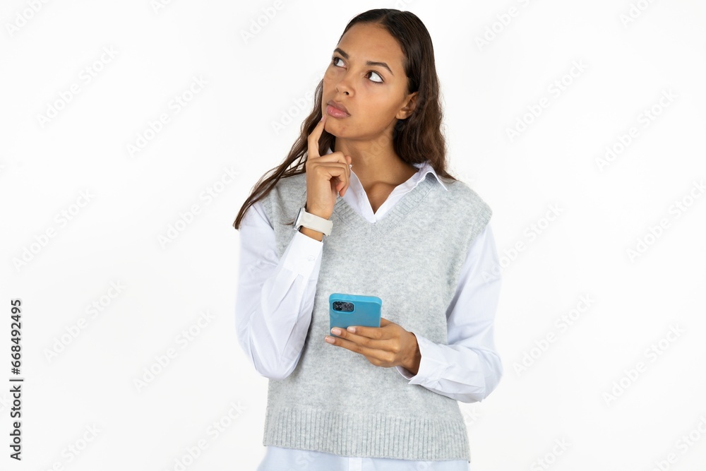 Young beautiful woman thinks deeply about something, uses modern mobile phone, tries to made up good message, keeps index finger near lips. Young beautiful woman thinks deeply about something, uses modern mobile phone, tries to made up good message, keeps index finger near lips.