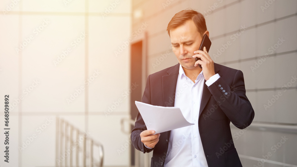 Concentrated mature businessman dressed in suit walks outdoors talking on phone. Adult concerned man talks with interest holding documents, sunlight Concentrated mature businessman dressed in suit walks outdoors talking on phone. Adult concerned man talks with interest holding documents, sunlight