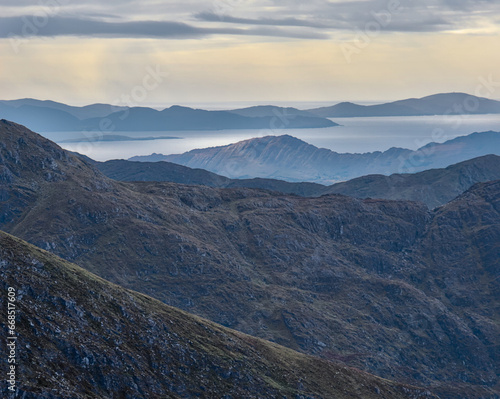A dramatic telephoto view of the Atlantic Ocean meets layers of steep mountain slopes lit by sunshine peeking from a cloudy sky in the distance. Mangerton County Kerry, Ireland