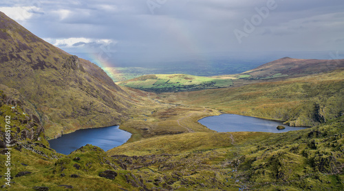 A broad panorama of two rainbows peeking through a cloudy sky to highlight a large valley surrounded by lush hills and steep mountains, and in it, a meandering river that forms two sparkling lakes