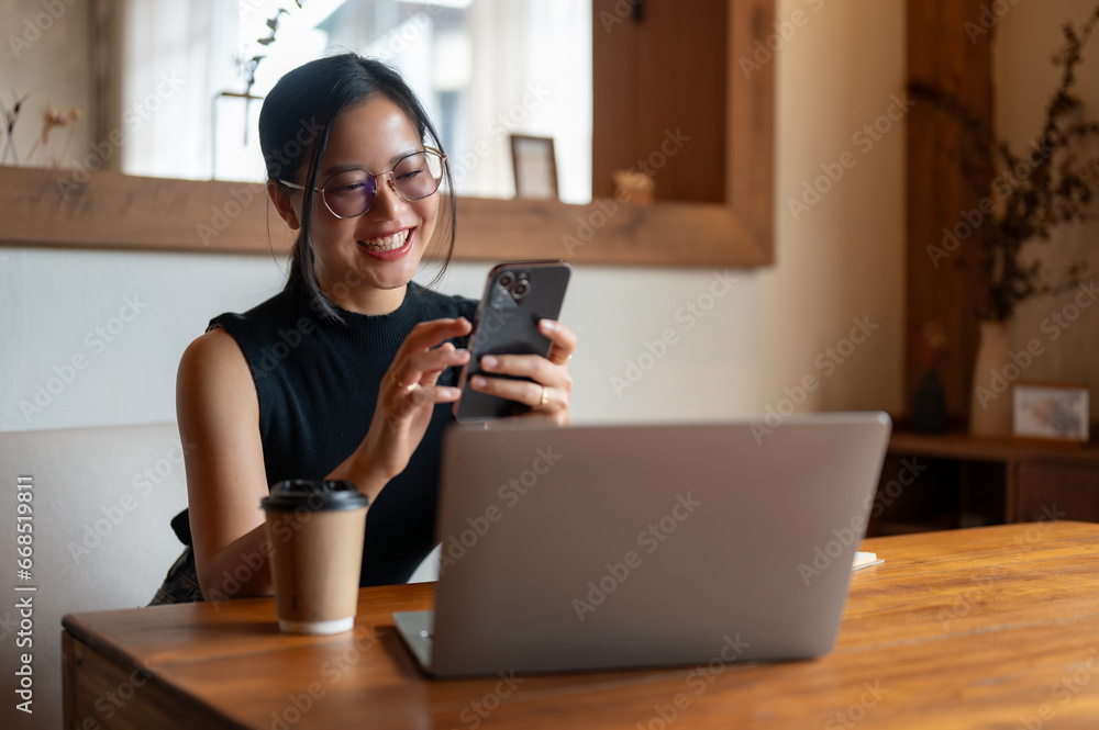 A cheerful Asian woman enjoys chatting with her friends on her phone while sitting in a coffee shop.