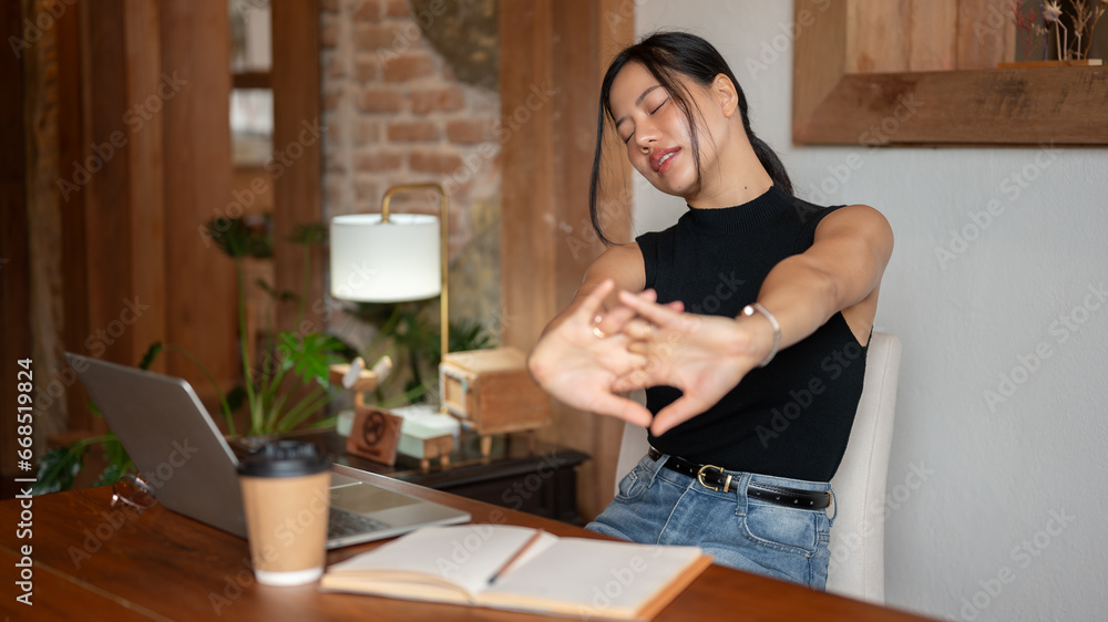 A tired Asian woman is stretching her arms while working on her tasks remotely at a coffee shop.