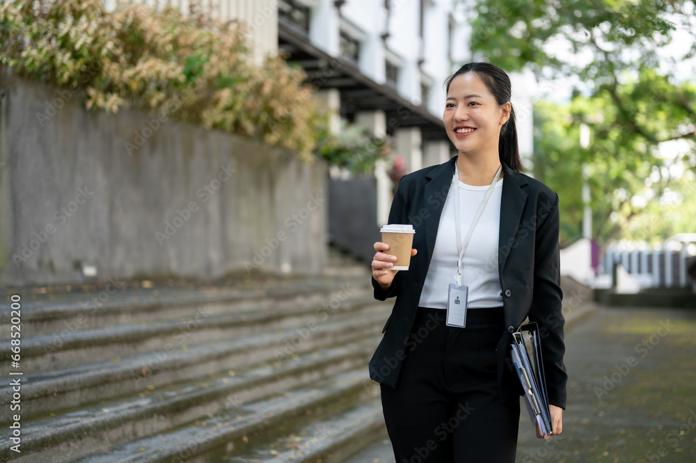 A cheerful businesswoman with a takeaway coffee cup and binders in her hands is walking in the city.