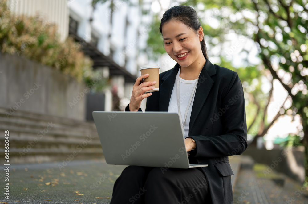 A happy businesswoman is having her morning coffee while working on her laptop on the stairs. A happy businesswoman is having her morning coffee while working on her laptop on the stairs.
