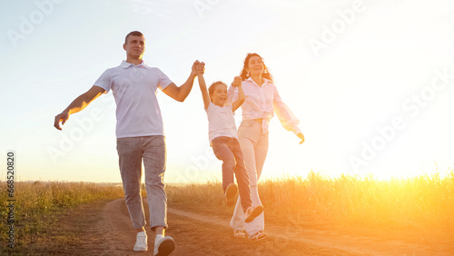 Parents lift daughter taking hands walking in field at sunset. Sun rays fall on happy family. Concept of positivity and spending time together, sunlight