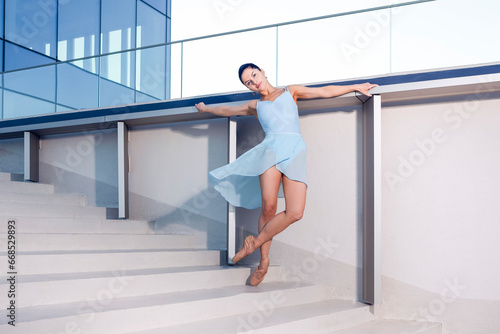 Slim Professional Caucasian Ballet Dancer in Blue Tutu Dreass Whie Posing Against White Wall In Leg Muscle Stretching Pose On Stairs In Stretching