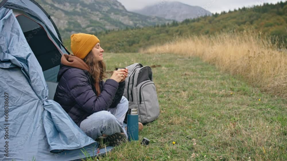 A young female traveler drinks hot tea from a thermos while sitting in a tent during her solo trip to the mountains. A young female traveler drinks hot tea from a thermos while sitting in a tent during her solo trip to the mountains.