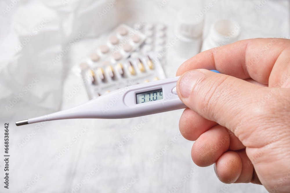 Hand holding a digital thermometer and pills on a white background.