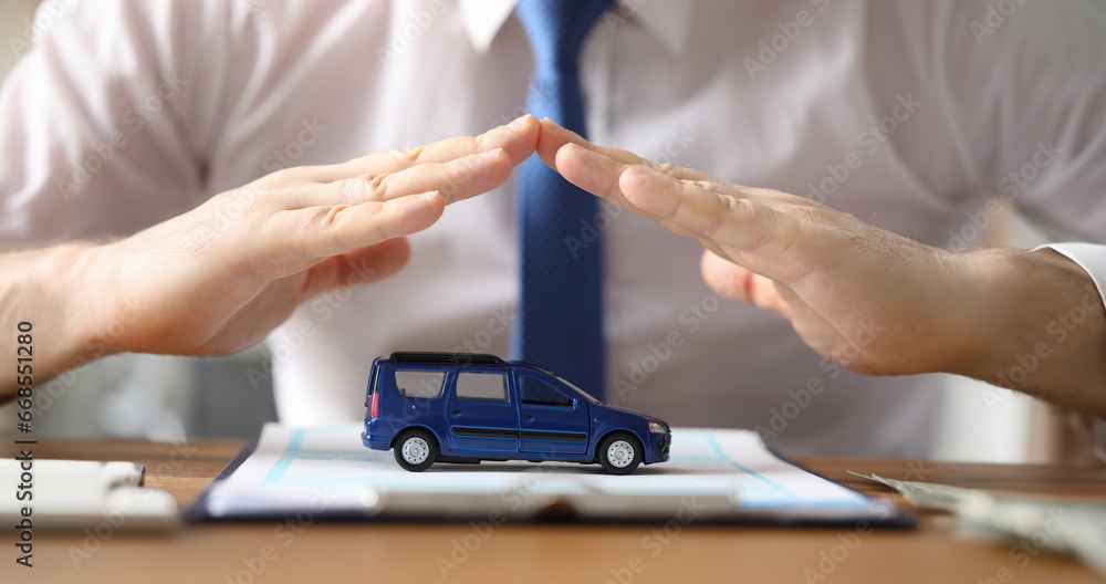 Close-up view of mans hands showing protection above blue car model. Businessman investing in transport. Presentable male in suit at office. Automobile insurance concept