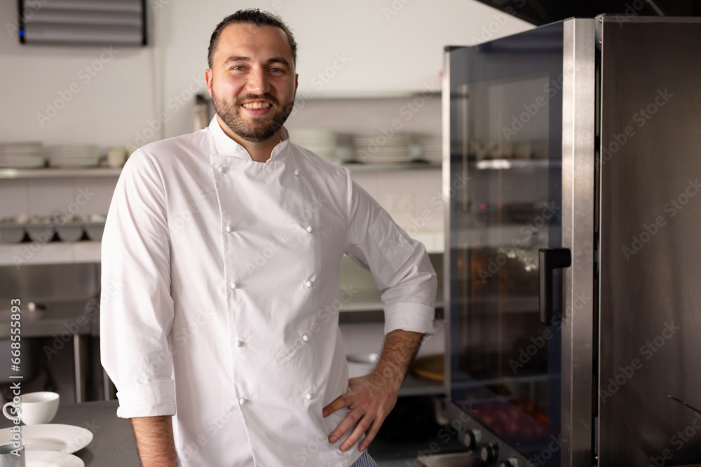 Portrait of smiling caucasian male chef with hand on hip standing in kitchen at cooking school Portrait of smiling caucasian male chef with hand on hip standing in kitchen at cooking school
