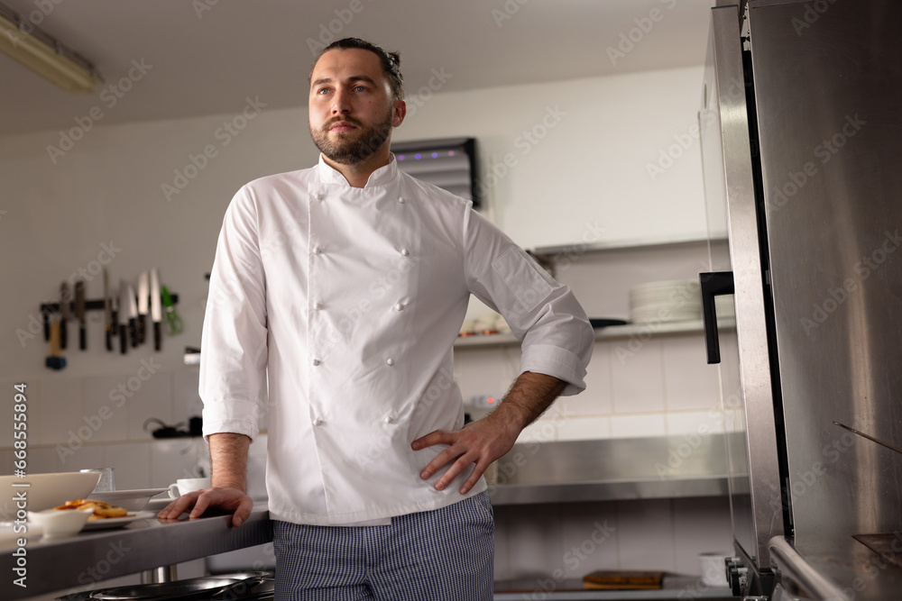 Thoughtful caucasian male chef with hand on hip standing in kitchen at cooking school Thoughtful caucasian male chef with hand on hip standing in kitchen at cooking school