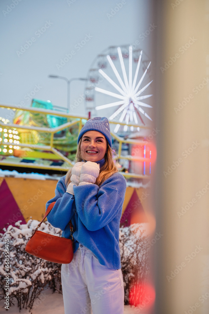 Cheerful and stylish woman, dressed in warm clothes, is having fun in a snowy winter amusement park