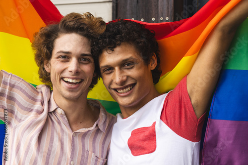 Portrait of happy diverse gay male couple holding lgbt flag and embracing at balcony