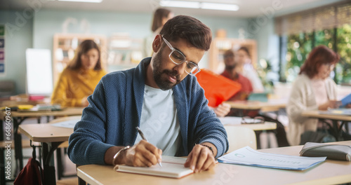 Portrait of a Handsome Indian Student Taking a Course in an International Adult Education Center. South Asian Man Wearing Glasses, Sitting Behind a Desk and Writing Down Notes in Notebook