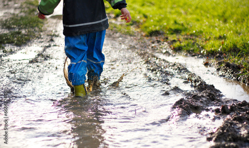 Lower body of a boy wearing blue waterproof pants and rubber boots walking in a muddy puddle during an autumn or spring walk. Outdoor fun in the rainy weather. Health and immunity of children concept