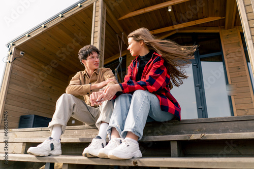 A young stylish couple in love sits together hugging on the steps of large wooden cottage and chats cheerfully © Guys Who Shoot