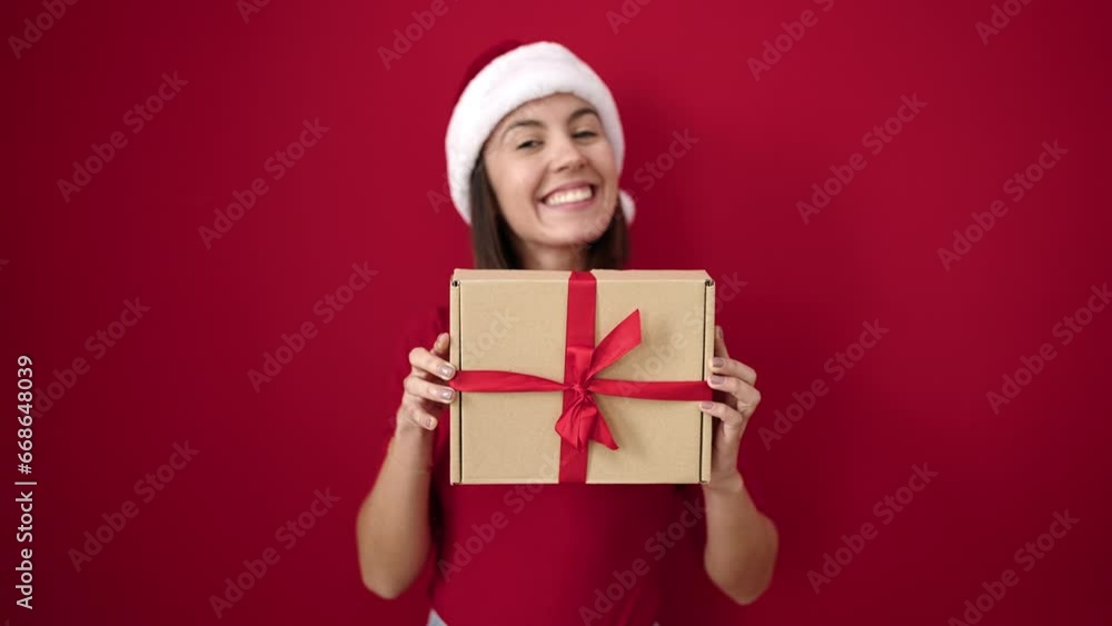 Young beautiful hispanic woman wearing christmas hat holding gift over isolated red background Young beautiful hispanic woman wearing christmas hat holding gift over isolated red background