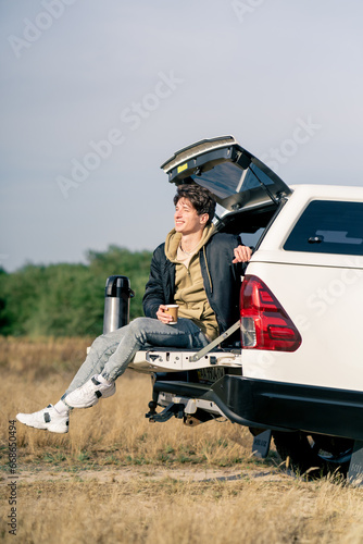 A pensive guy sits in the open trunk of his car with a thermos and glass of tea outdoors in field