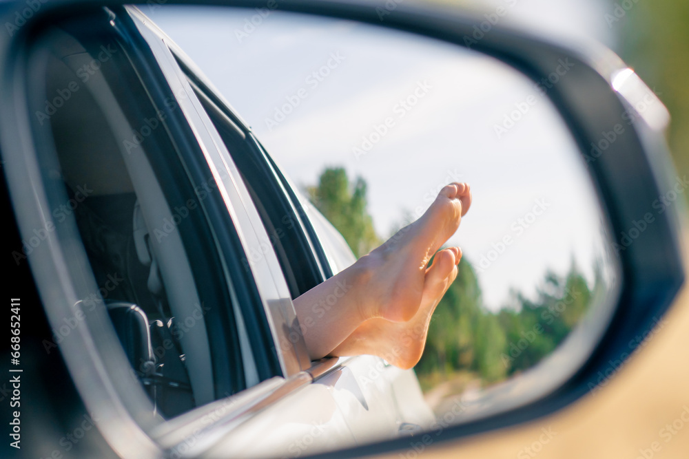 Naklejka premium In the side mirror of a car the reflection of a woman’s legs stretched out of window onto the street while driving through field