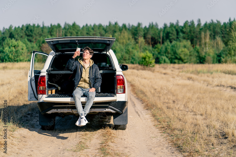 The car is standing on the road in the forest-steppe A young guy sits in the open trunk with phone and gets connection