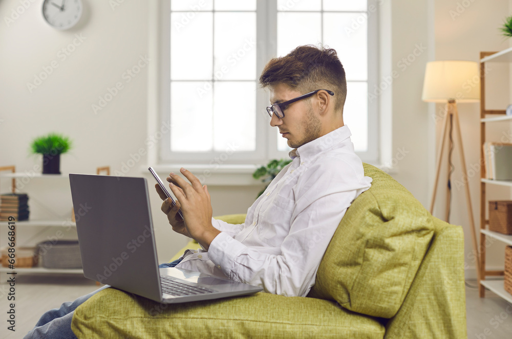 Young man sitting on couch at home and using cellphone. Profile view of serious businessman in glasses sitting on sofa with laptop, looking at screen of mobile phone, dialing number or sending message Young man sitting on couch at home and using cellphone. Profile view of serious businessman in glasses sitting on sofa with laptop, looking at screen of mobile phone, dialing number or sending message