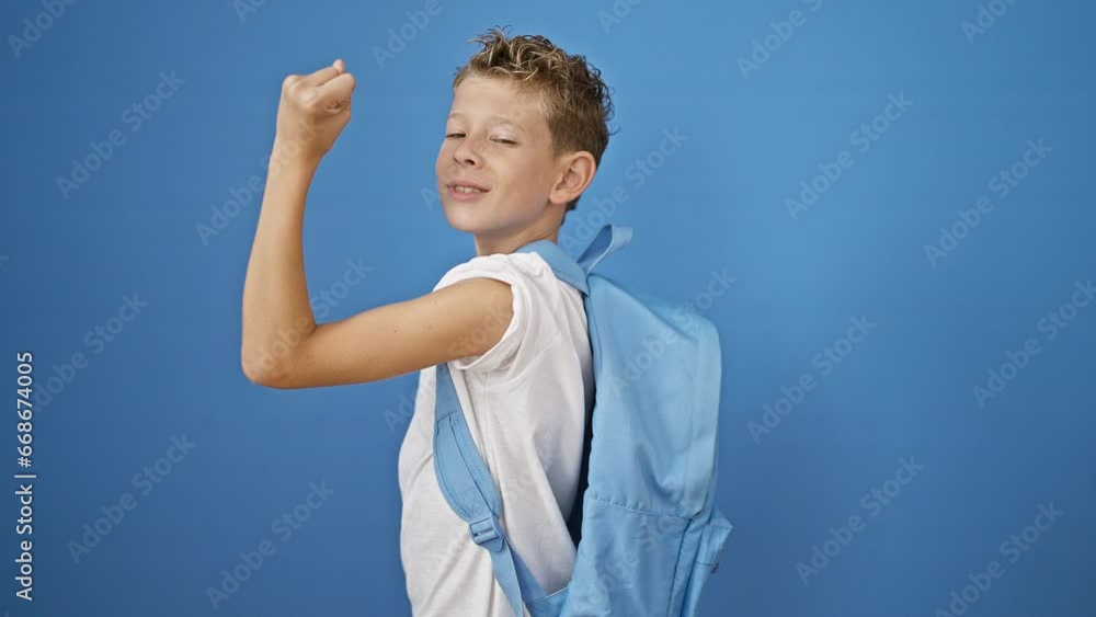 Adorable blond boy student standing isolated on blue background, confidently showing off strong arm gesture while enjoying his learning journey Adorable blond boy student standing isolated on blue background, confidently showing off strong arm gesture while enjoying his learning journey