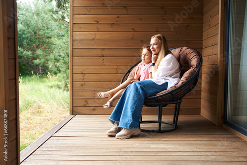 Smiling daughter and attractive mother sitting in armchair on large porch
