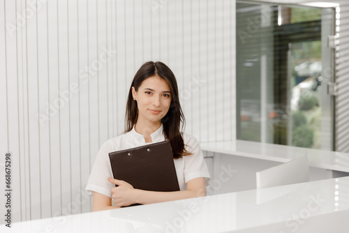 In front of the camera beautiful medical assistant posing to the camera at reception desk she smiling cute holding a map with documents in hands