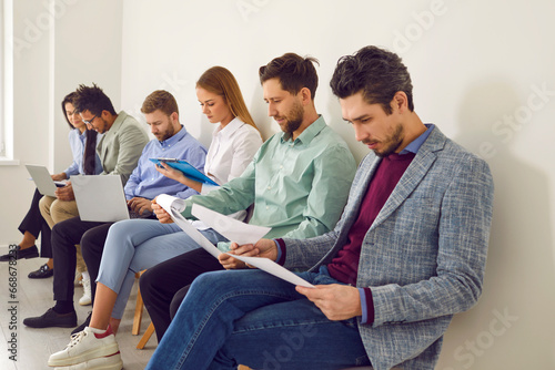 Job applicants sitting in queue waiting for job interview in office. Group of diverse people reading resumes preparing for job interview. Human resources, employment, recruitment