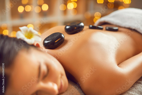 Close up shot of young woman having spa hot stone massage. Relaxed pretty girl lying on couch with black basalt stones along spine. Beauty treatment therapy, body care concept