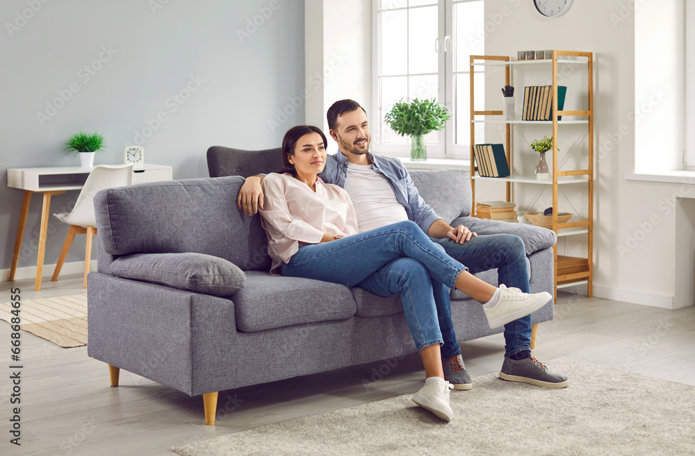 Happy relaxed young family couple sitting on sofa at home. Smiling husband and wife watching television, enjoying carefree leisure weekend time. Family lifestyle, recreation concept Happy relaxed young family couple sitting on sofa at home. Smiling husband and wife watching television, enjoying carefree leisure weekend time. Family lifestyle, recreation concept