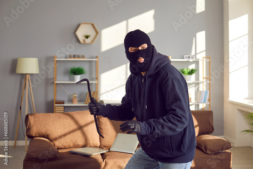 Burglar with crowbar in the house. Robber man wearing black jacket, balaclava mask and gloves breaks into apartment. Sneaky thief walking on tiptoes and looking around for valuable stuff he can take photo